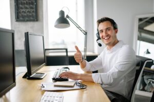 Happy man in office wearing headset, giving a thumbs up while typing at a computer.